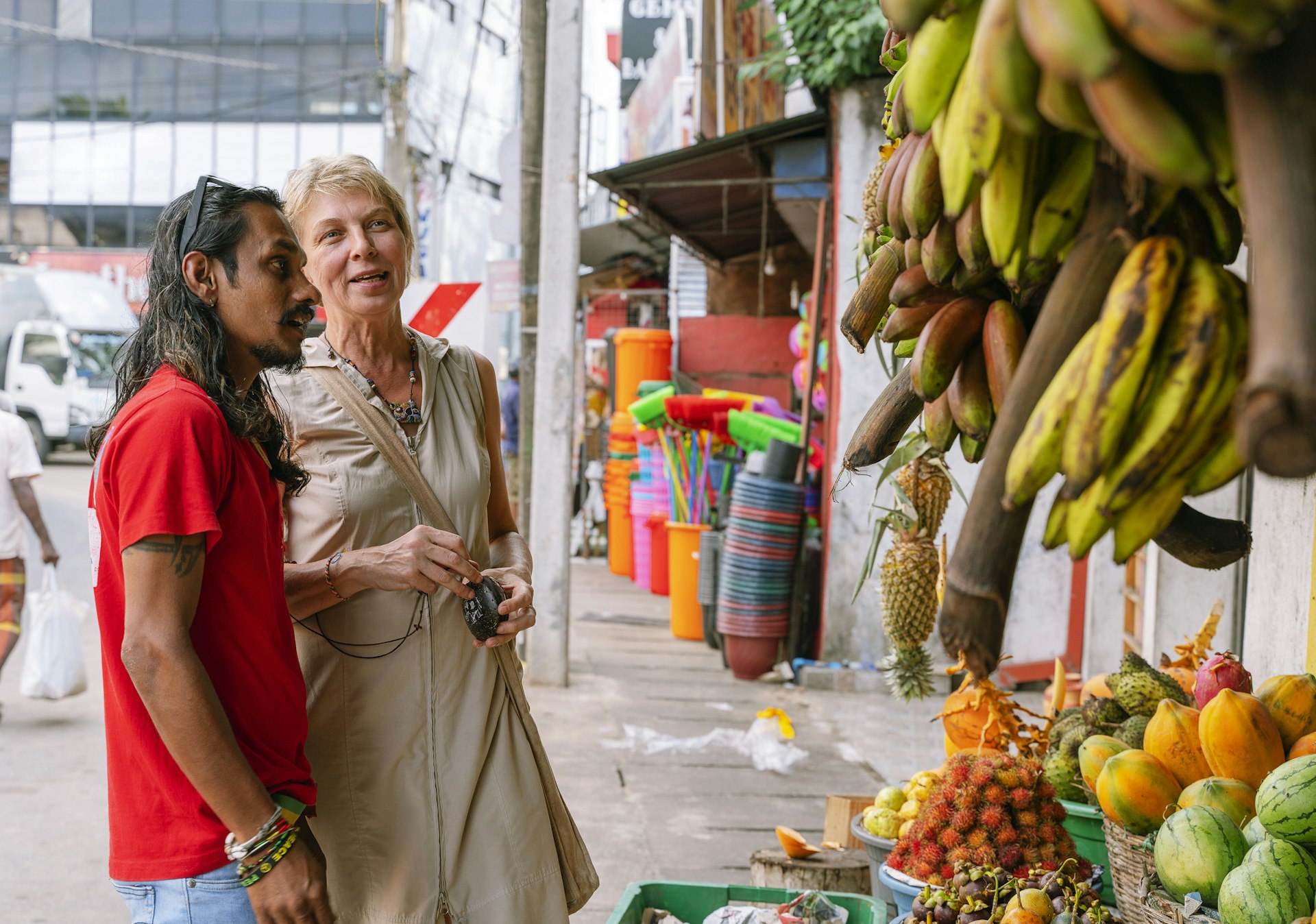 A young Sri Lankan man and a white older lady stand at a fruit stall in a city